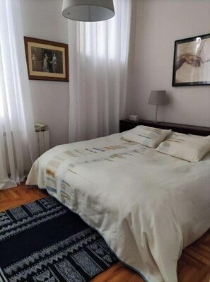 A neatly arranged bedroom with a bed, a bedside lamp, a framed picture on the wall, and a window with curtains.