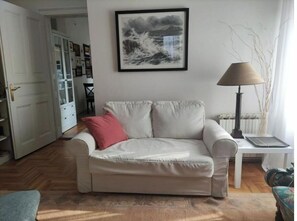 A living room with a beige sofa, a red pillow, a framed artwork of a seascape, a lamp, and a radiator.