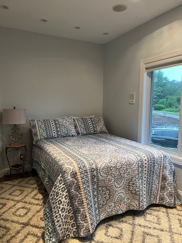 A bedroom with a patterned bedspread, a bedside table with a lamp, and a window with a view of greenery.