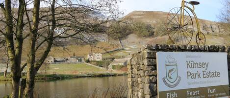 A sign for Kilnsey Park Estate with a view of a lake, stone wall, and a bicycle.