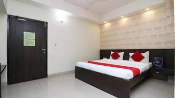 Quarto com cama de madeira escura, roupa de cama branca e almofadas vermelhas. Há um ventilador de teto, uma porta com uma placa e uma parede com padrão.