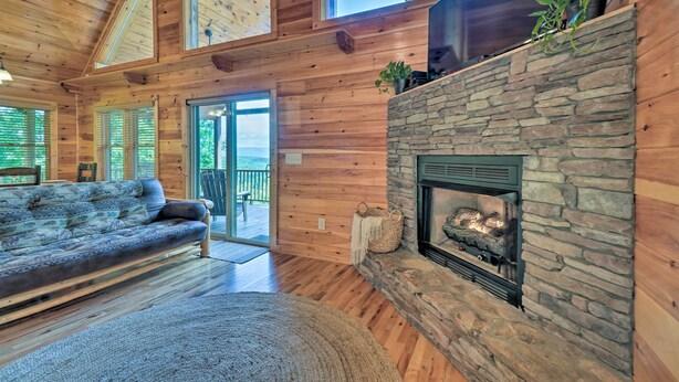 A cozy cabin interior with a stone fireplace, wooden walls, a sofa, and a large window. - ‘Hiwassee Hideaway' Cabin w/ Mountain Views! (Murphy)