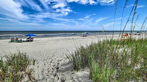 A beach with people under umbrellas, a lifeguard tower, and tall grass in the foreground. - Myrtle Beach Condo w/ On-Site Pool & Beach Access (Myrtle Beach)