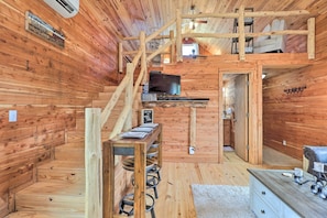 Interior de una cabaña de madera con escalera de madera, televisor, cartel de bienvenida y una pequeña zona de kitchenette.