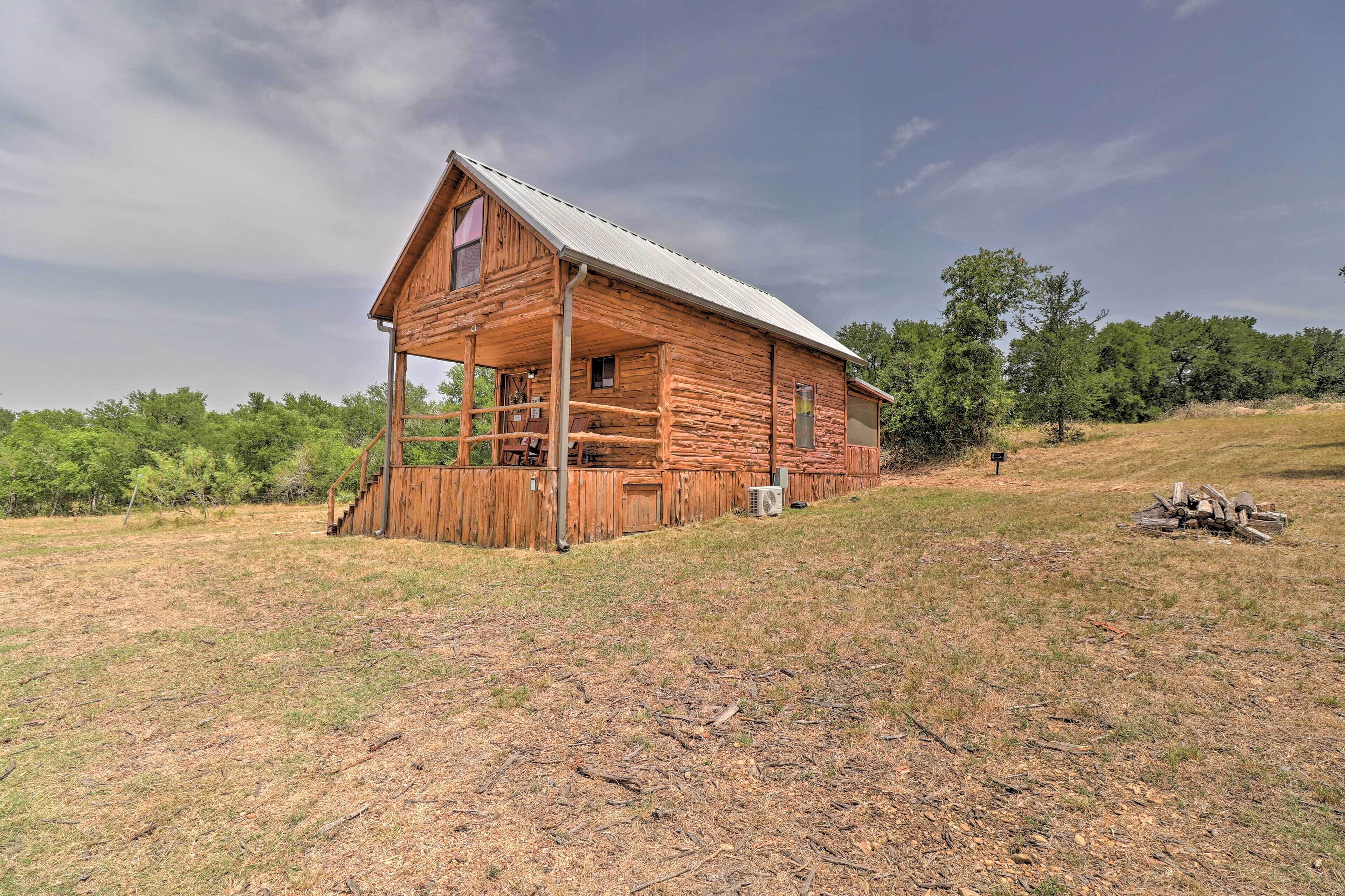 A rustic log cabin with a porch, situated on a grassy hill with trees in the background.