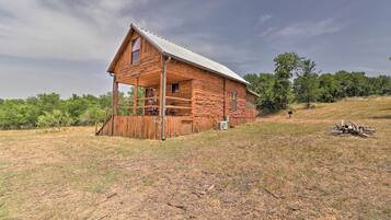 A rustic log cabin with a porch, situated on a grassy hill with trees in the background.