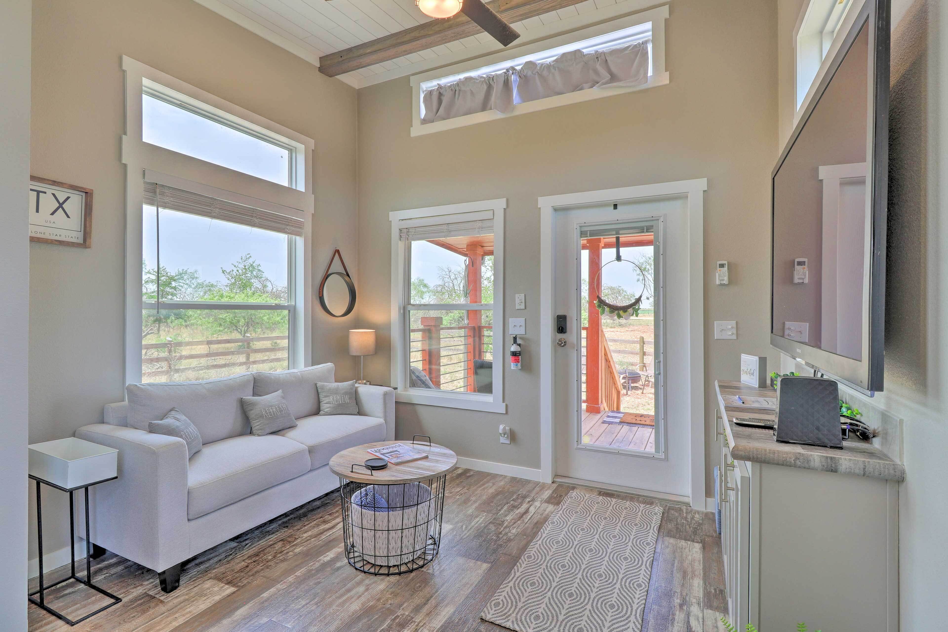A modern living room with a white sofa, a round coffee table, and a TV mounted on the wall. There are large windows and a door leading to an outdoor area.