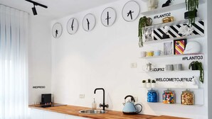 A modern kitchen with a wooden countertop, a kettle, and a sink. There are wall-mounted clocks and shelves with decorative items and plants. - Yehuda Market Suite - One Bedroom Apartment, Sleeps 4 (Jerusalem)