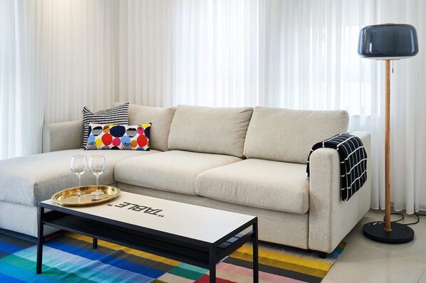 Un salon avec un canapé beige, des coussins colorés, une table basse avec un plateau et des verres, et un lampadaire.