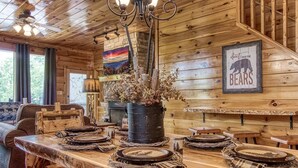 A rustic wooden interior with a dining table set for four, a chandelier, and a bear-themed sign.