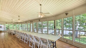 Una larga mesa con sillas blancas en una habitación amplia con suelo de madera y ventanas grandes.