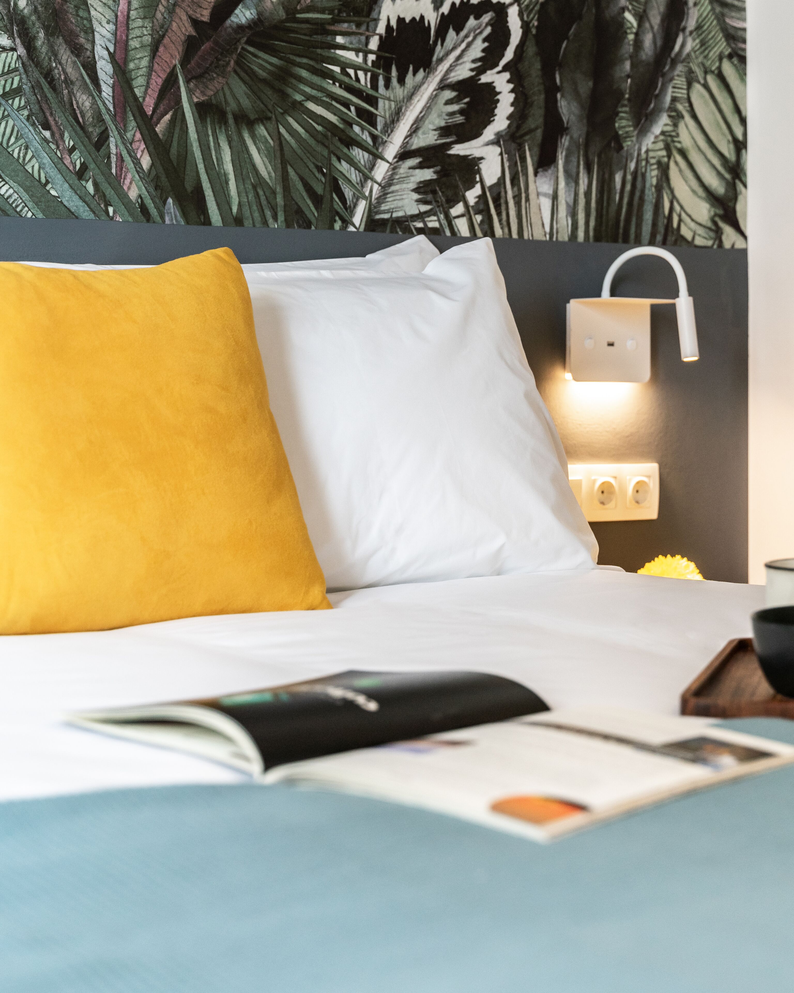 Ein ordentlich bezogenes Bett mit weißen und gelben Kissen, eine Zeitschrift auf dem Bett und eine Steckdose mit Mehrfachsteckdosenleiste.