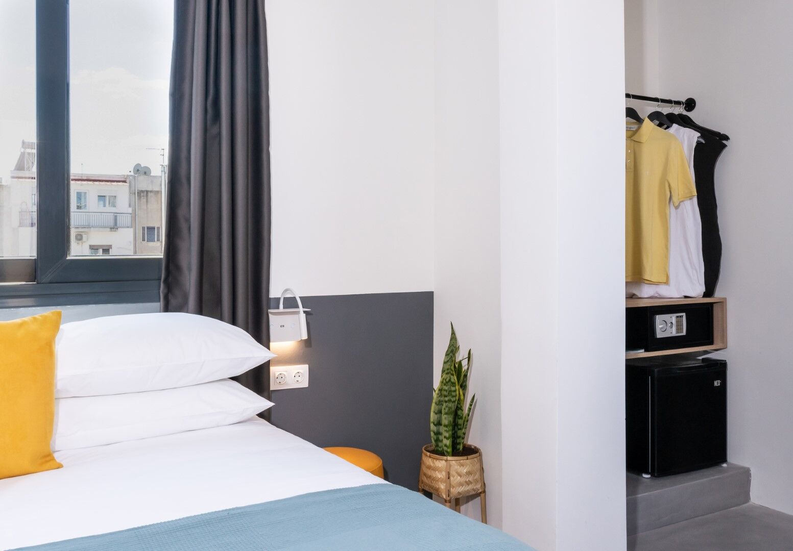 A modern hotel room with a bed, a bedside table with a lamp, a small potted plant, and a coat rack with hanging clothes.
