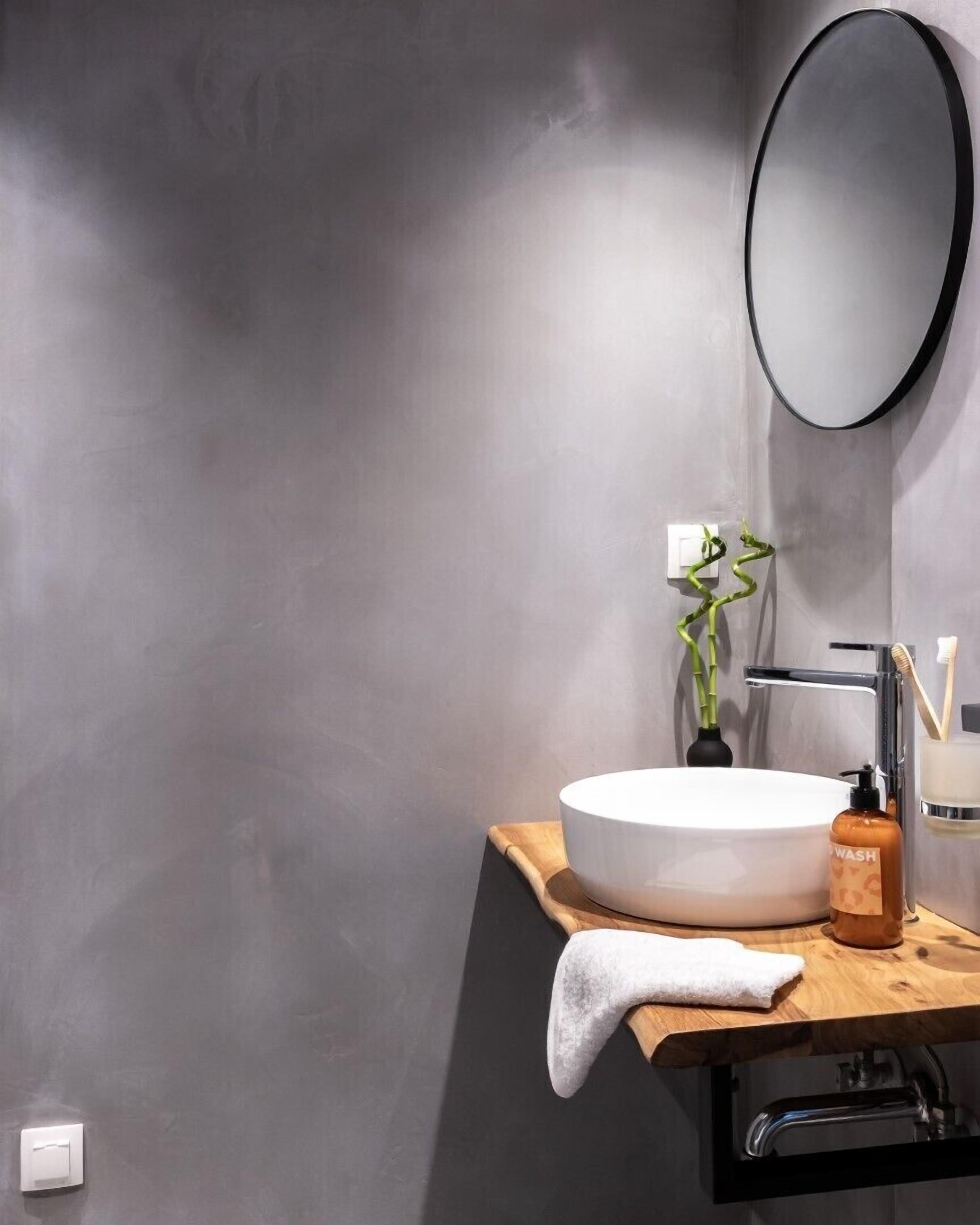 Ein modernes Badezimmer mit einem runden weißen Waschbecken, einem schwarzen Wasserhahn, einer Holz-Arbeitsplatte, einem Spiegel, einer grünen Pflanze, einem Seifenspender und einem Handtuch.