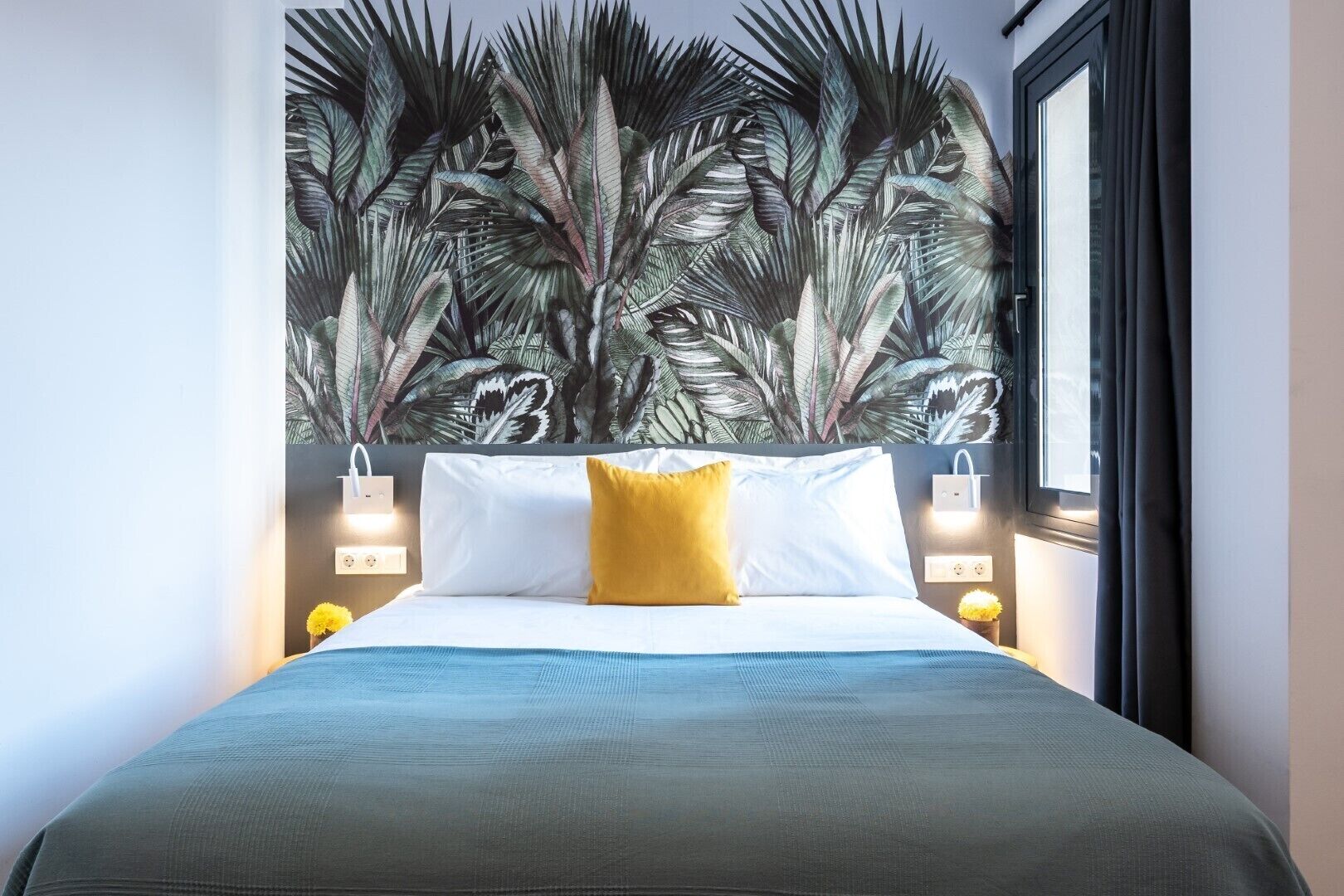 A bedroom with a bed, a yellow pillow, a large tropical leaf mural on the wall, and bedside lamps.