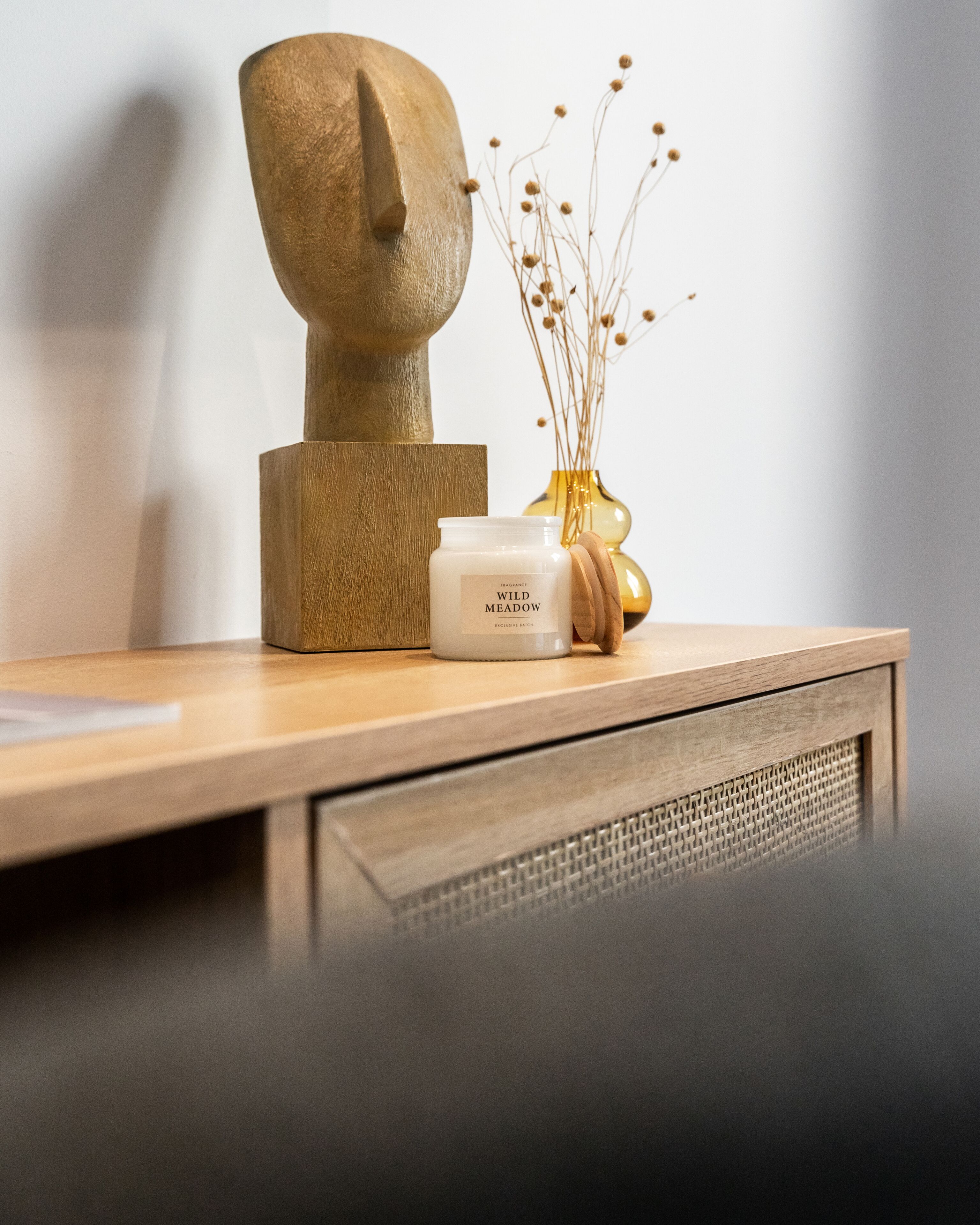 A wooden table with a white candle labeled "Wild Meadow" and a vase with dried flowers.