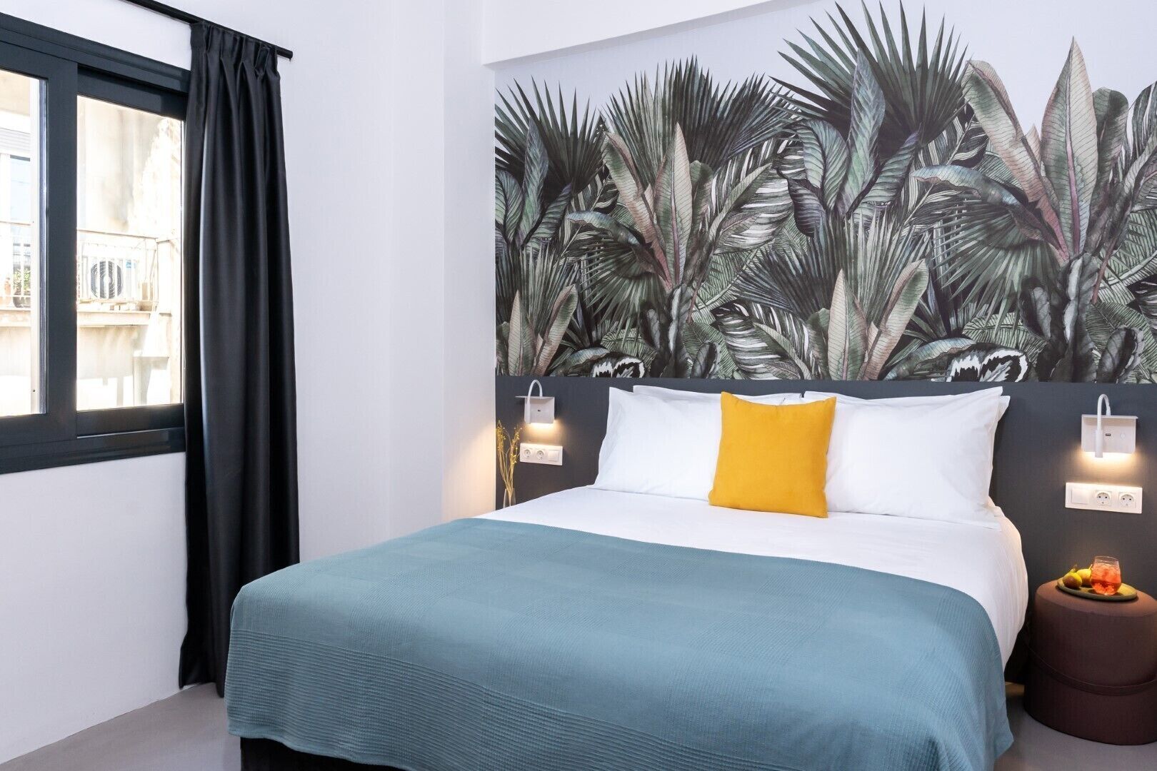 A bedroom with a bed, a wall mural of tropical leaves, a window with black curtains, and a small round table with fruit.