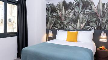 A bedroom with a bed, a wall mural of tropical leaves, a window with black curtains, and a small round table with fruit.