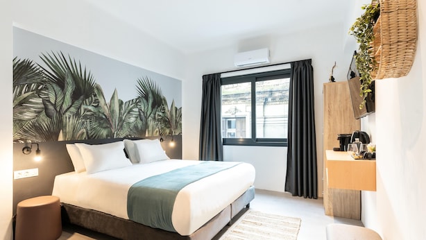Une chambre d’hôtel moderne dotée d’un grand lit, d’un téléviseur fixé au mur, d’une table de chevet avec une lampe, d’une fenêtre avec des rideaux et offrant une vue sur le paysage urbain.