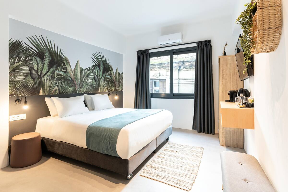 A modern hotel room with a large bed, a wall-mounted TV, a bedside table with a lamp, a window with curtains, and a view of the cityscape.
