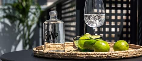 A table with a bottle, a glass, and limes on a woven mat, set outdoors with a building in the background.
