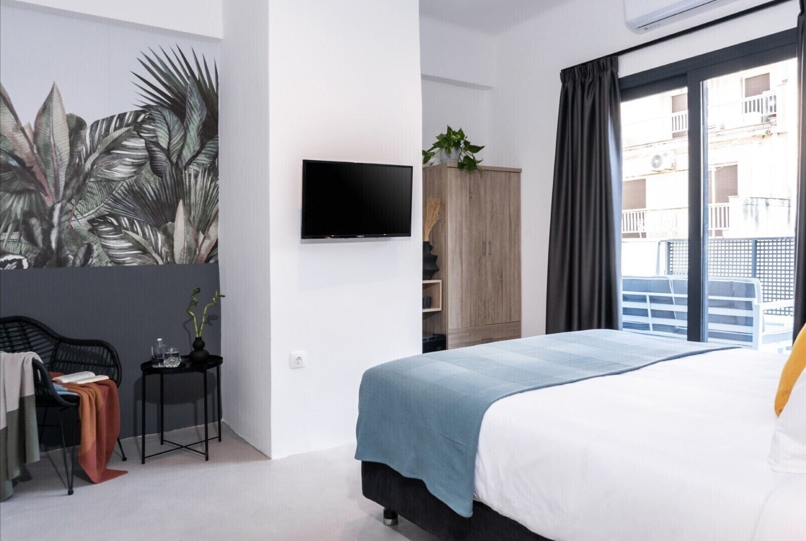 A modern hotel room with a large bed, a flat-screen TV, a small table with a plant, and a view of the city through the window.