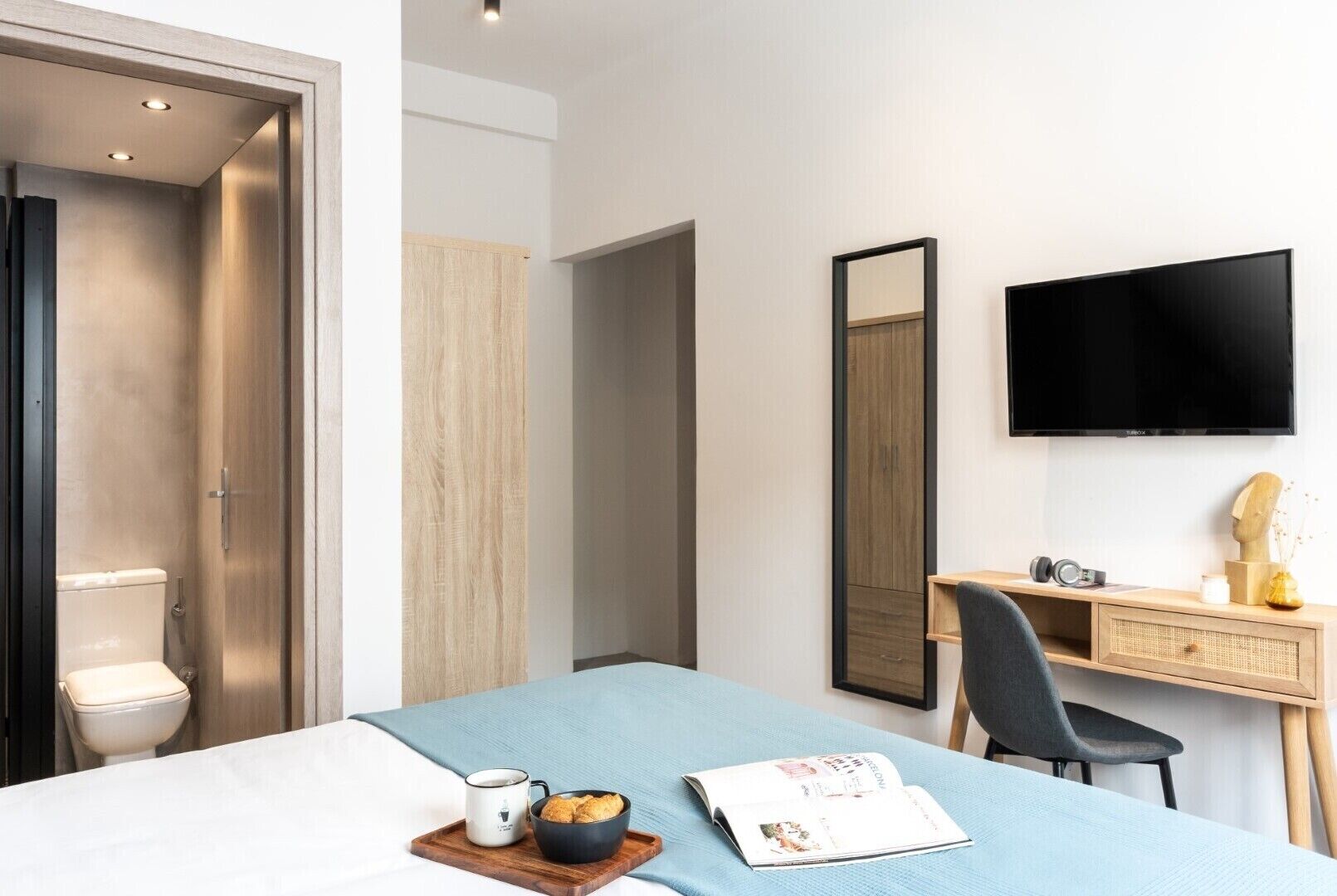 A modern hotel room with a bed, a desk, a chair, a TV, and a bathroom.