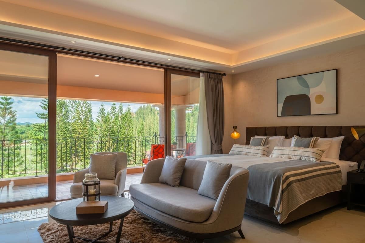 A modern bedroom with a large bed, a sofa, a small table, and a view of a balcony with trees.