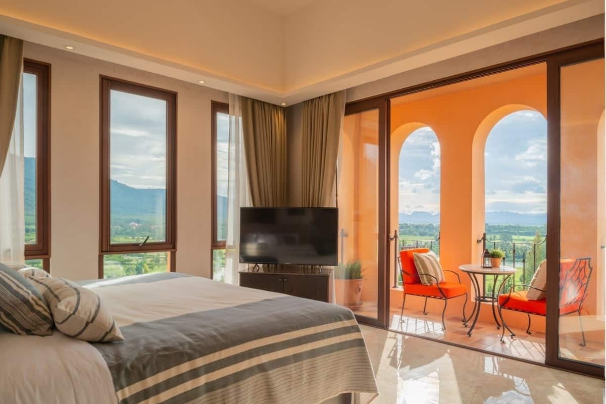 A bedroom with a large bed, a TV, and a balcony with red chairs.