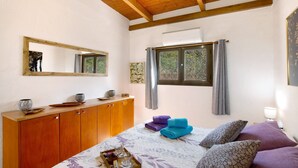 Ein Schlafzimmer mit Holzboden, einem Bett mit violetten und blauen Handtüchern, einer Holzkommode, einem Spiegel und einem Fenster mit Vorhängen.