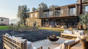 A modern outdoor seating area with wooden furniture and a fire pit, overlooking a stone building with a balcony.