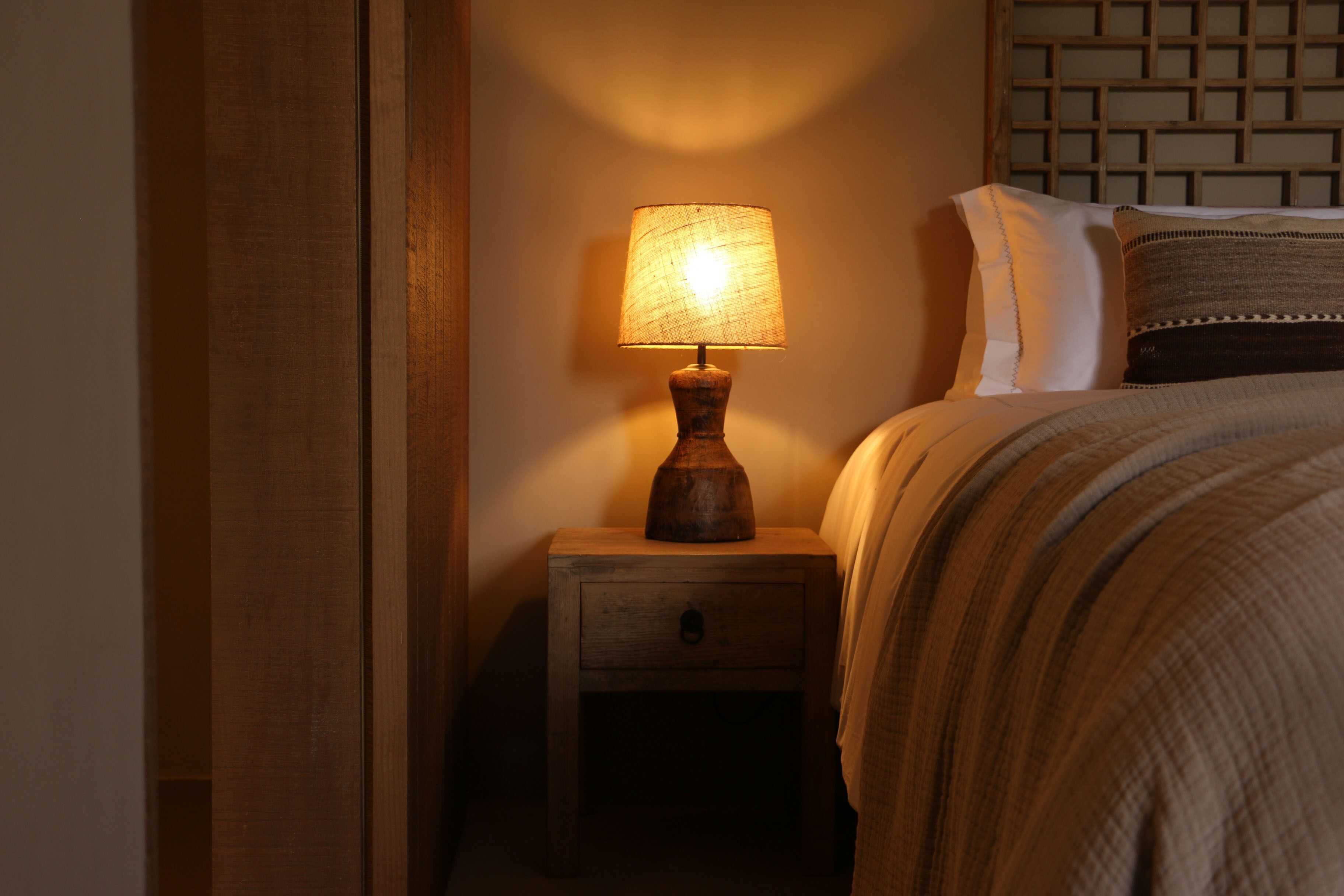 A wooden bedside table with a lamp, a bed with a striped blanket, and a headboard with a grid pattern.