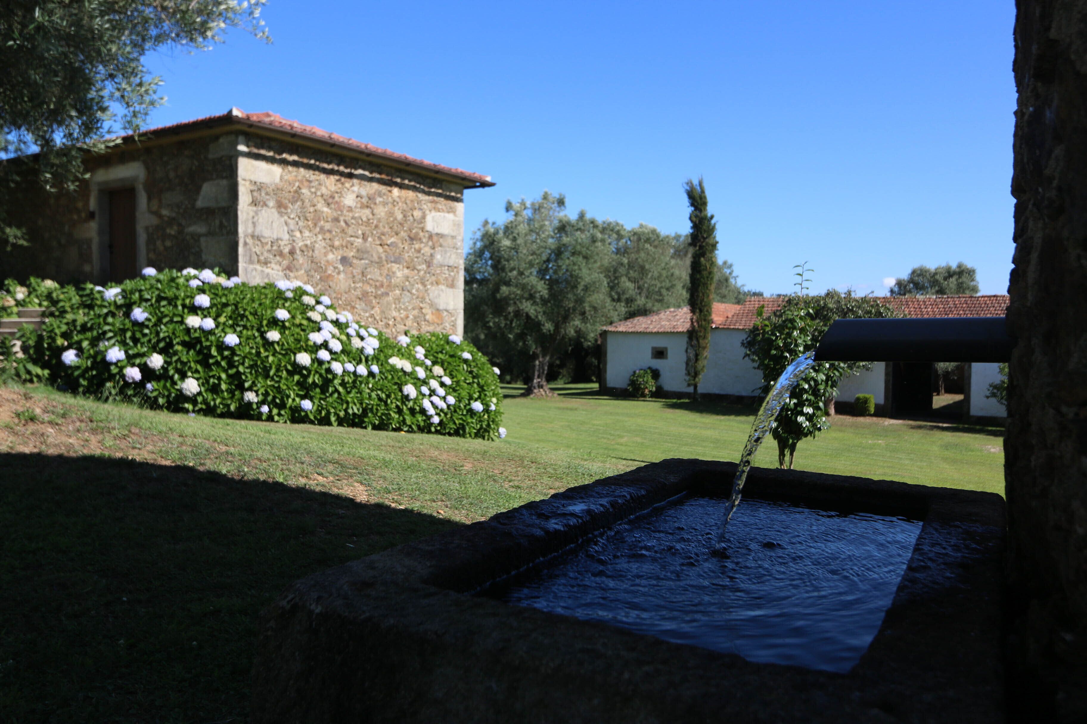 Un bâtiment en pierre avec un toit recouvert de tuiles rouges, une petite fontaine et un jardin agrémenté de fleurs blanches.