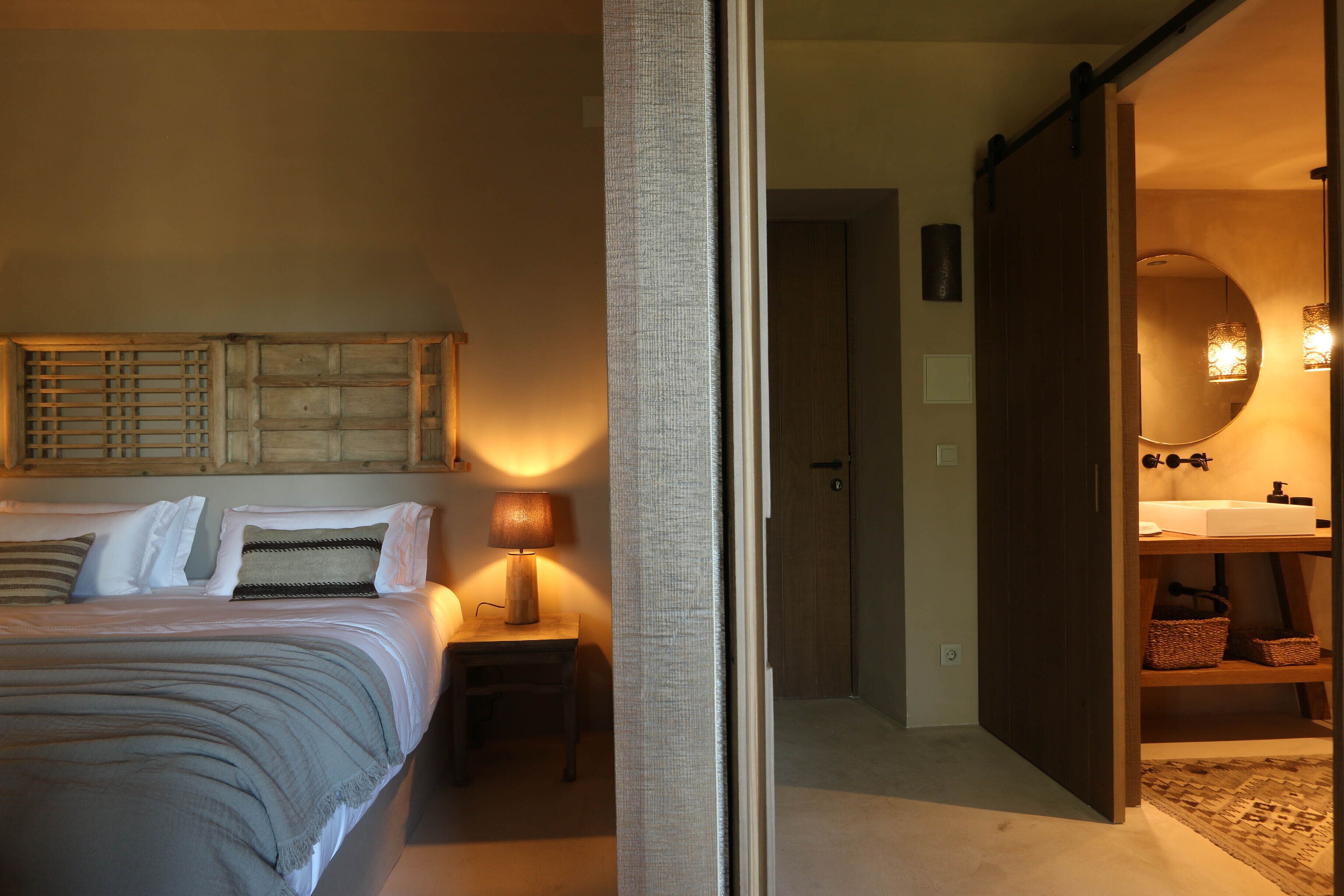 Une chambre à coucher comprenant un lit, une table de chevet, une lampe et donnant sur une salle de bain équipée d’un miroir et d’étagères.