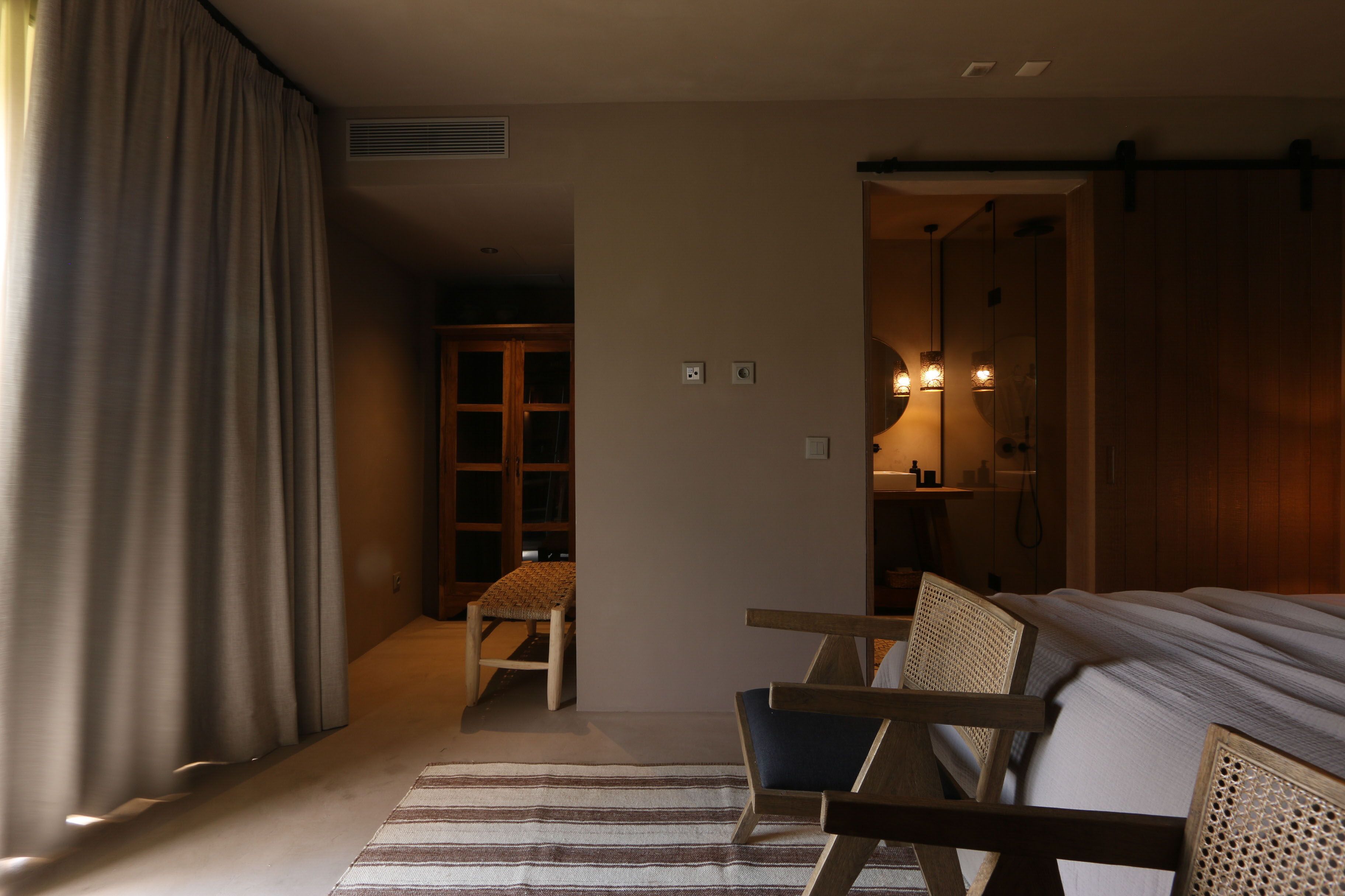 Habitación de hotel con cama, dos sillas de madera, alfombra a rayas, armario y un pequeño rincón de descanso con una silla y una mesa.