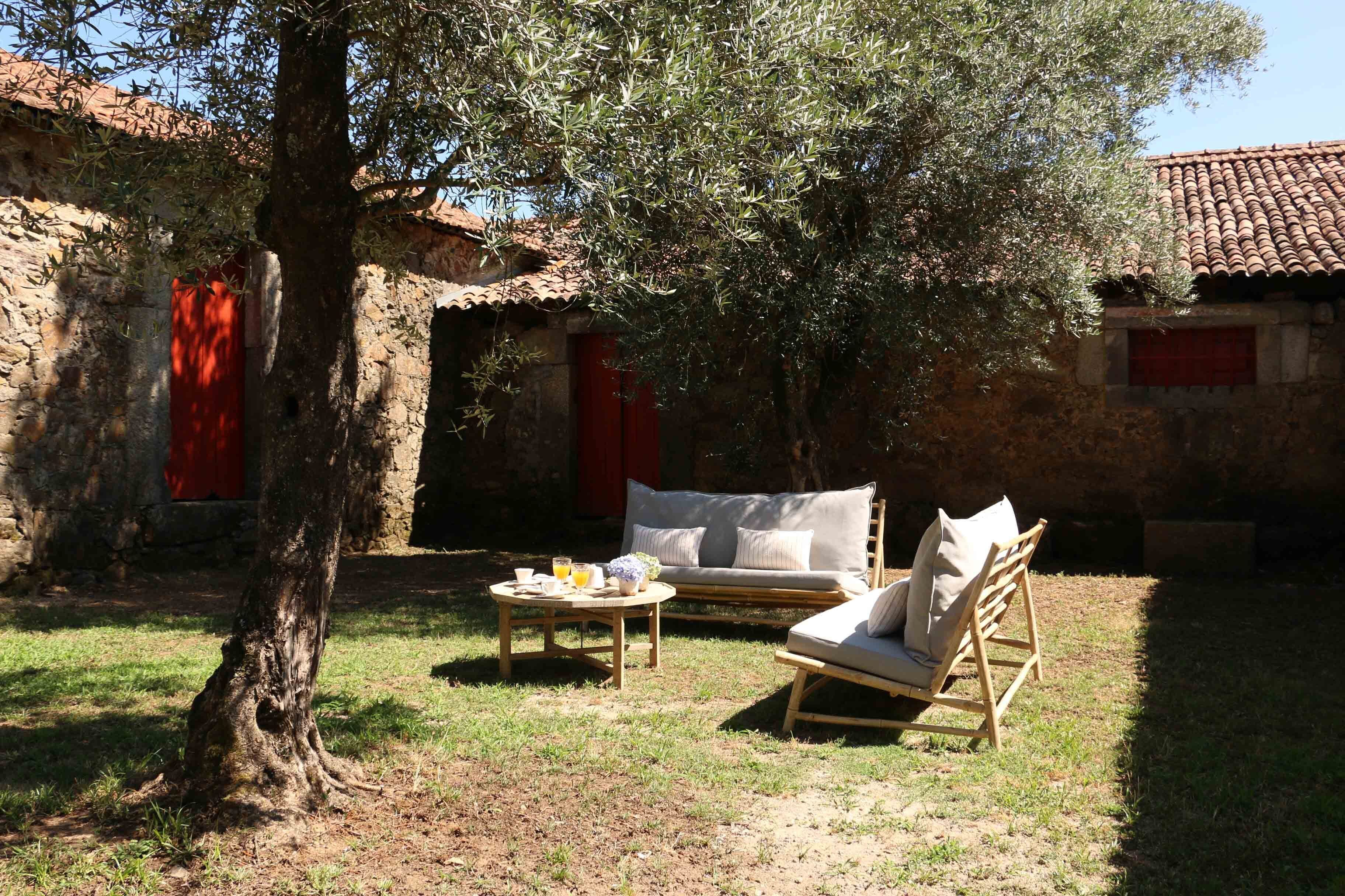 Un área de estar al aire libre de estilo rústico con una mesa de madera, dos sillas con cojines y una puerta roja al fondo.