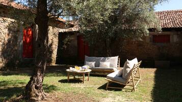 Un área de estar al aire libre de estilo rústico con una mesa de madera, dos sillas con cojines y una puerta roja al fondo.