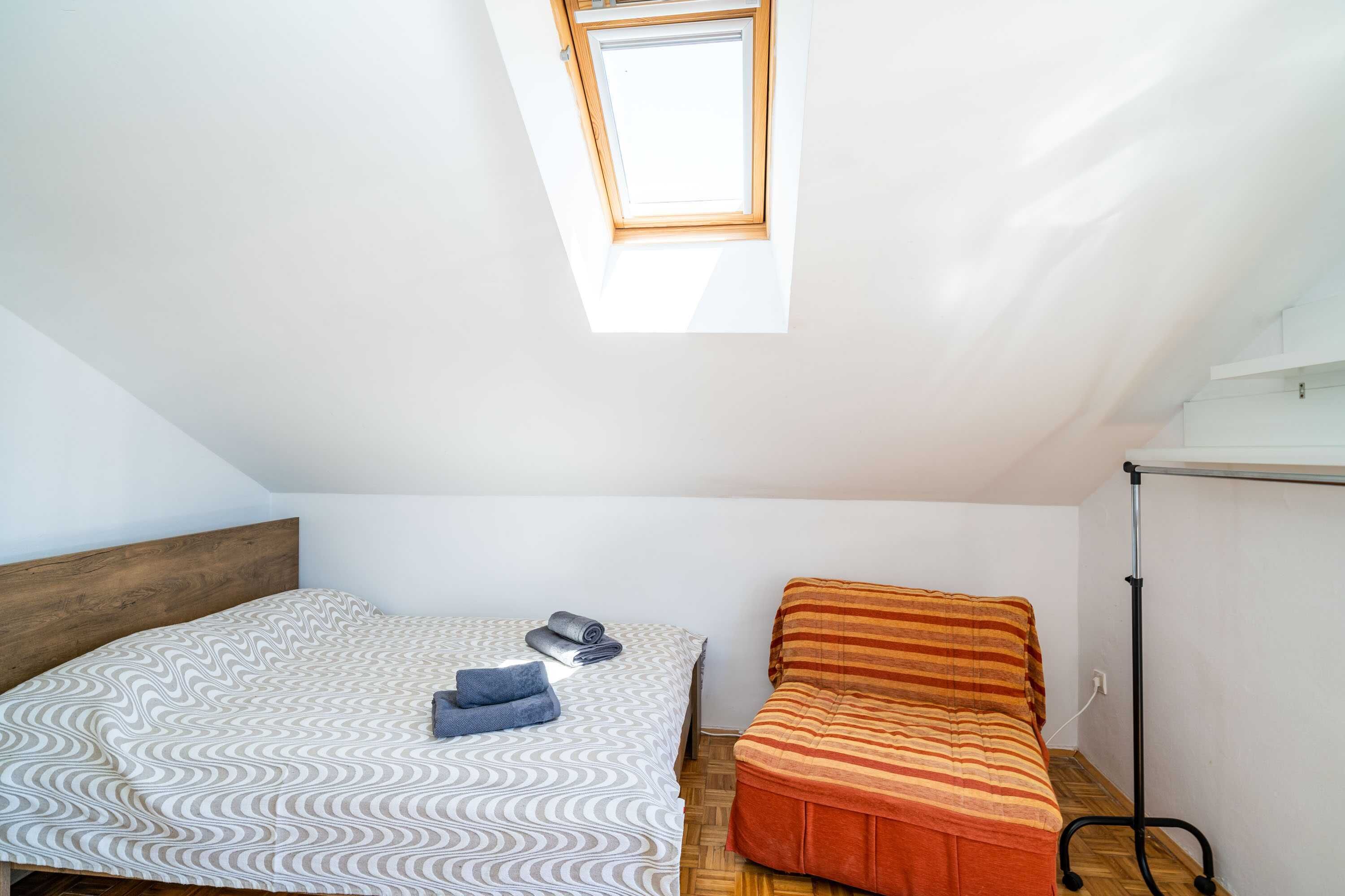 A small, bright room with a sloped ceiling, a skylight, a bed with a patterned bedspread, a bench with a striped cushion, and a black floor lamp.