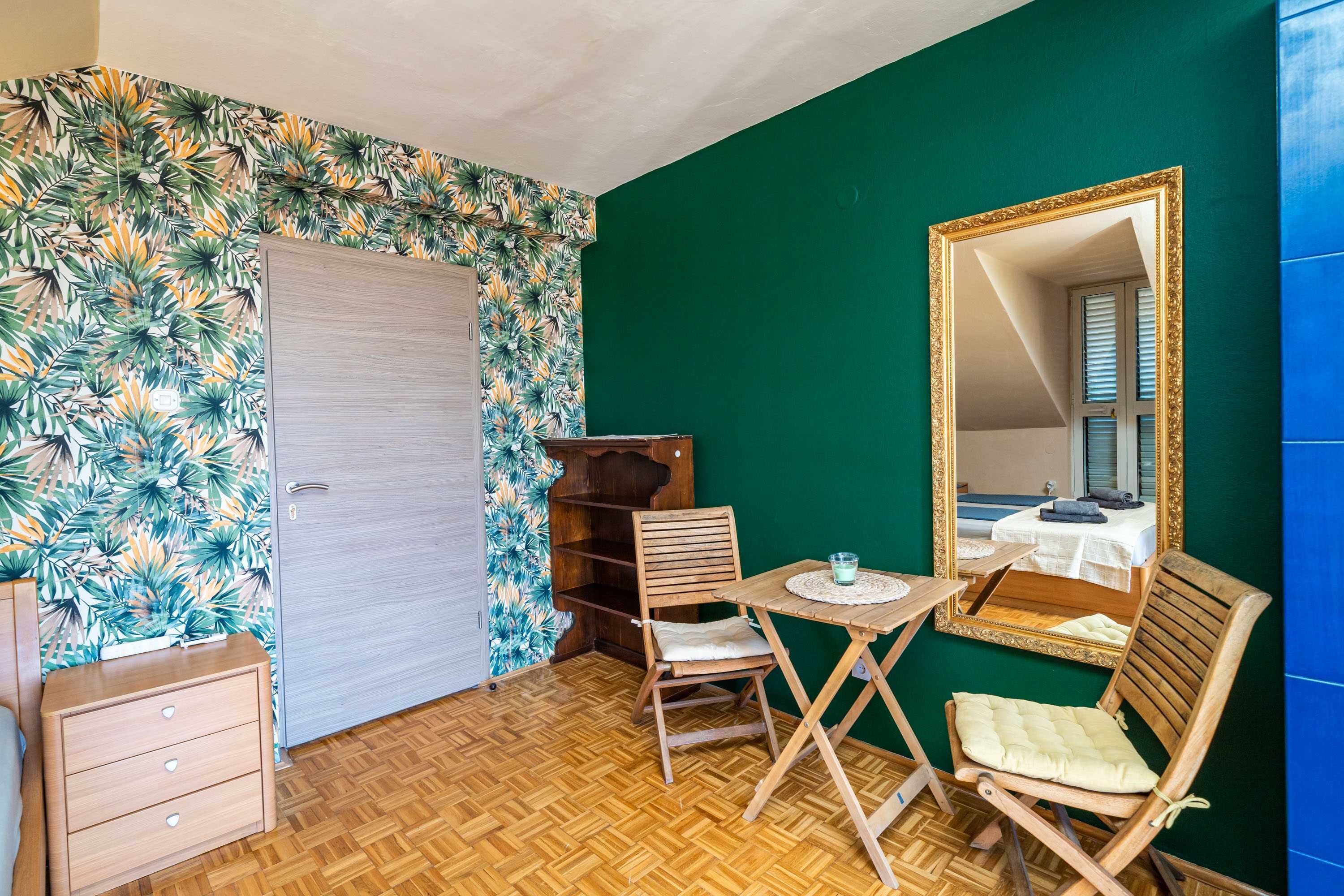 Una habitación con piso de madera, una silla de madera, una mesita con un vaso, un armario de madera y un espejo con marco dorado.