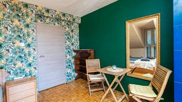 Una habitación con piso de madera, una silla de madera, una mesita con un vaso, un armario de madera y un espejo con marco dorado.