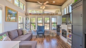 A spacious living room with a gray sofa, a blue armchair, and a fireplace. There are large windows and a ceiling fan. - Upscale Tiny Home - Boho-Chic Austin Getaway! (Austin)