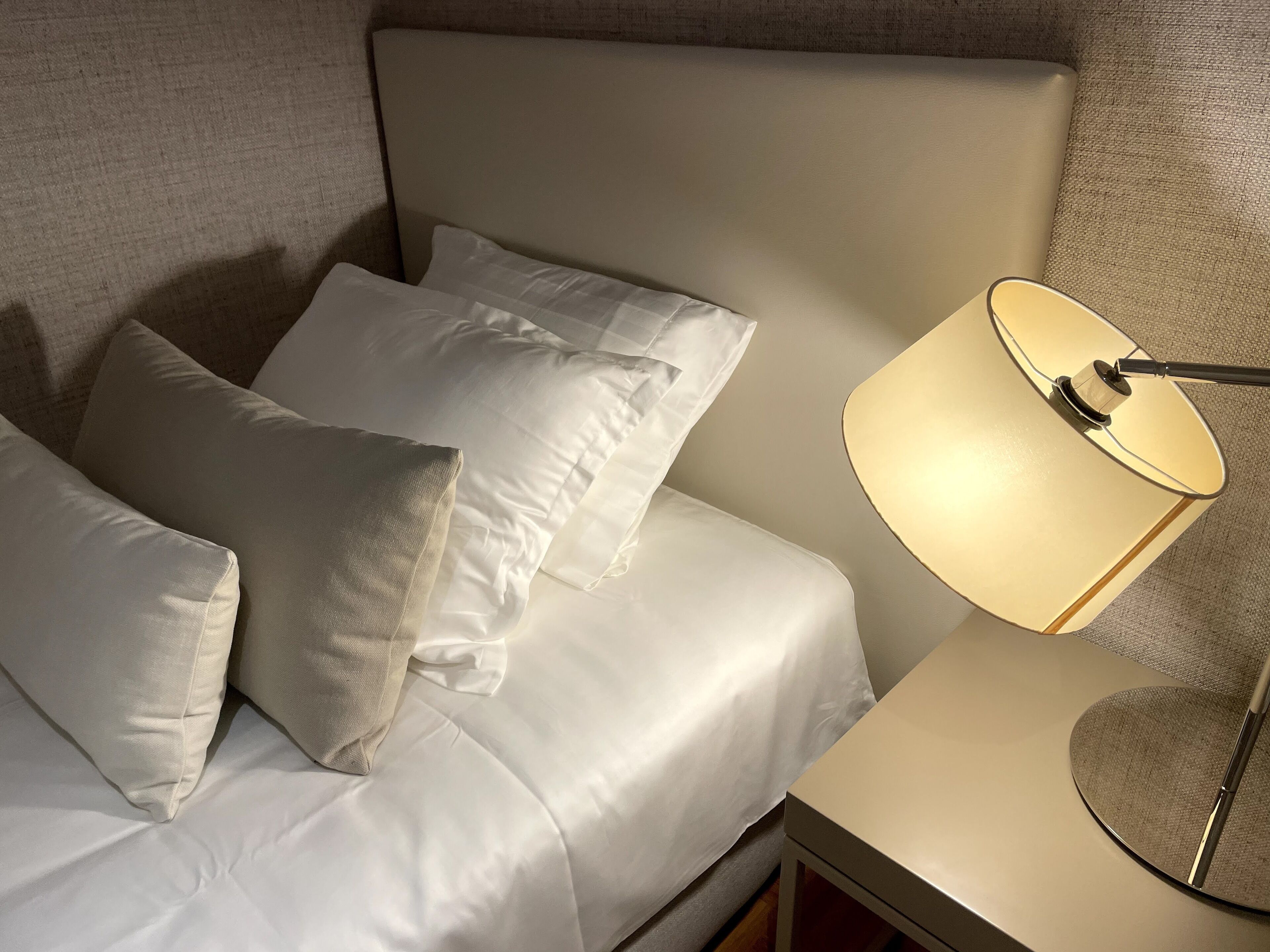 Una cama con sábanas y almohadas blancas, una lámpara de noche y un cabecero.