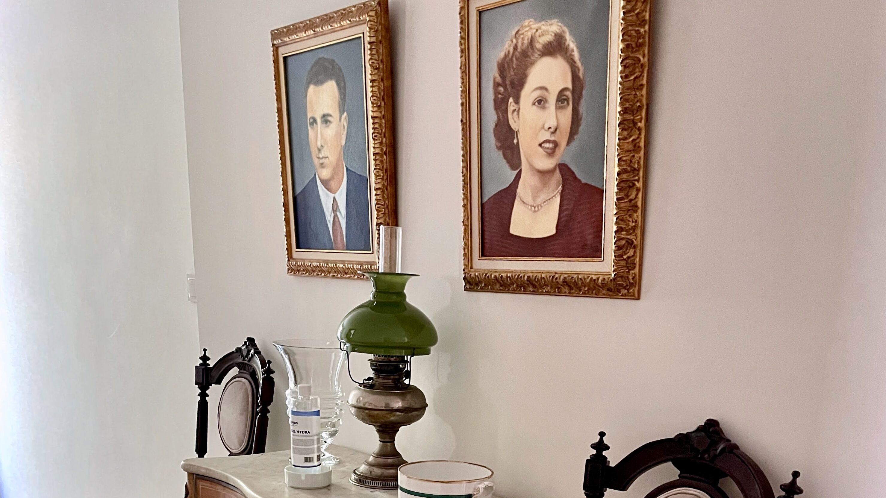 A room with two framed portraits on the wall, a green lamp on a table, a water bottle, and a cup.