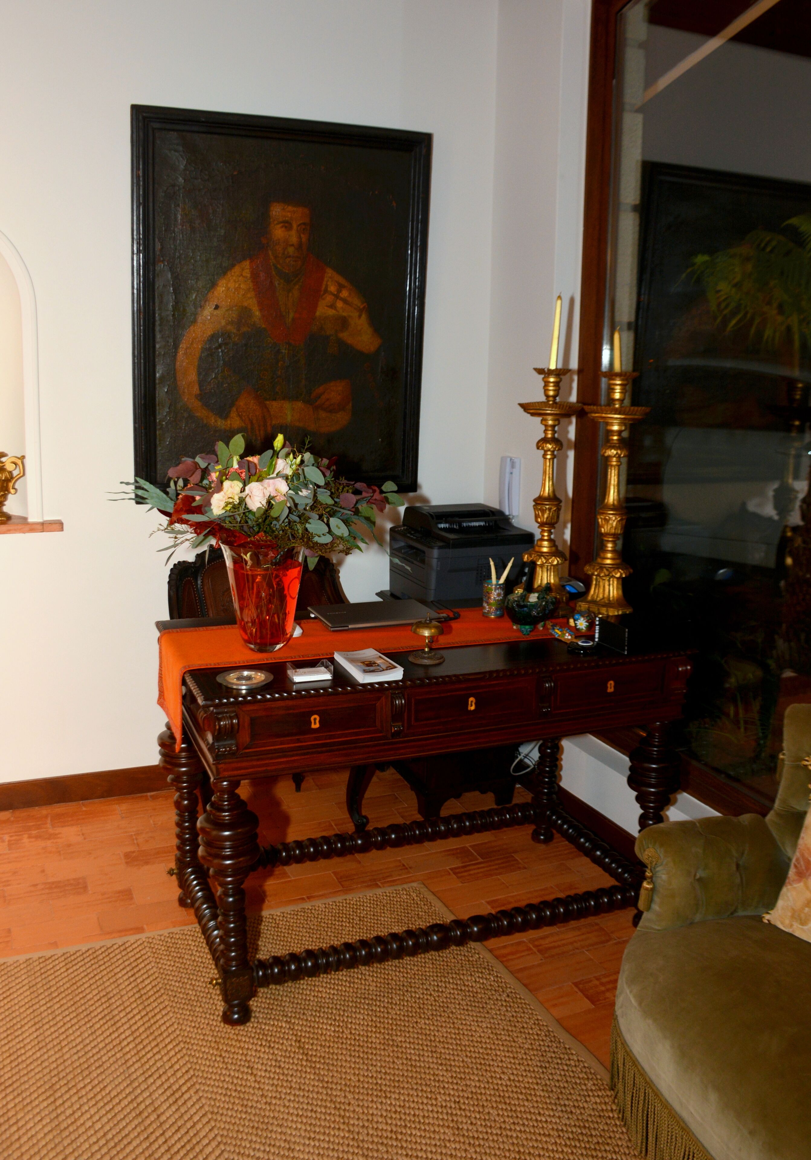 A wooden desk with a vase of flowers, a lamp, and a framed painting.