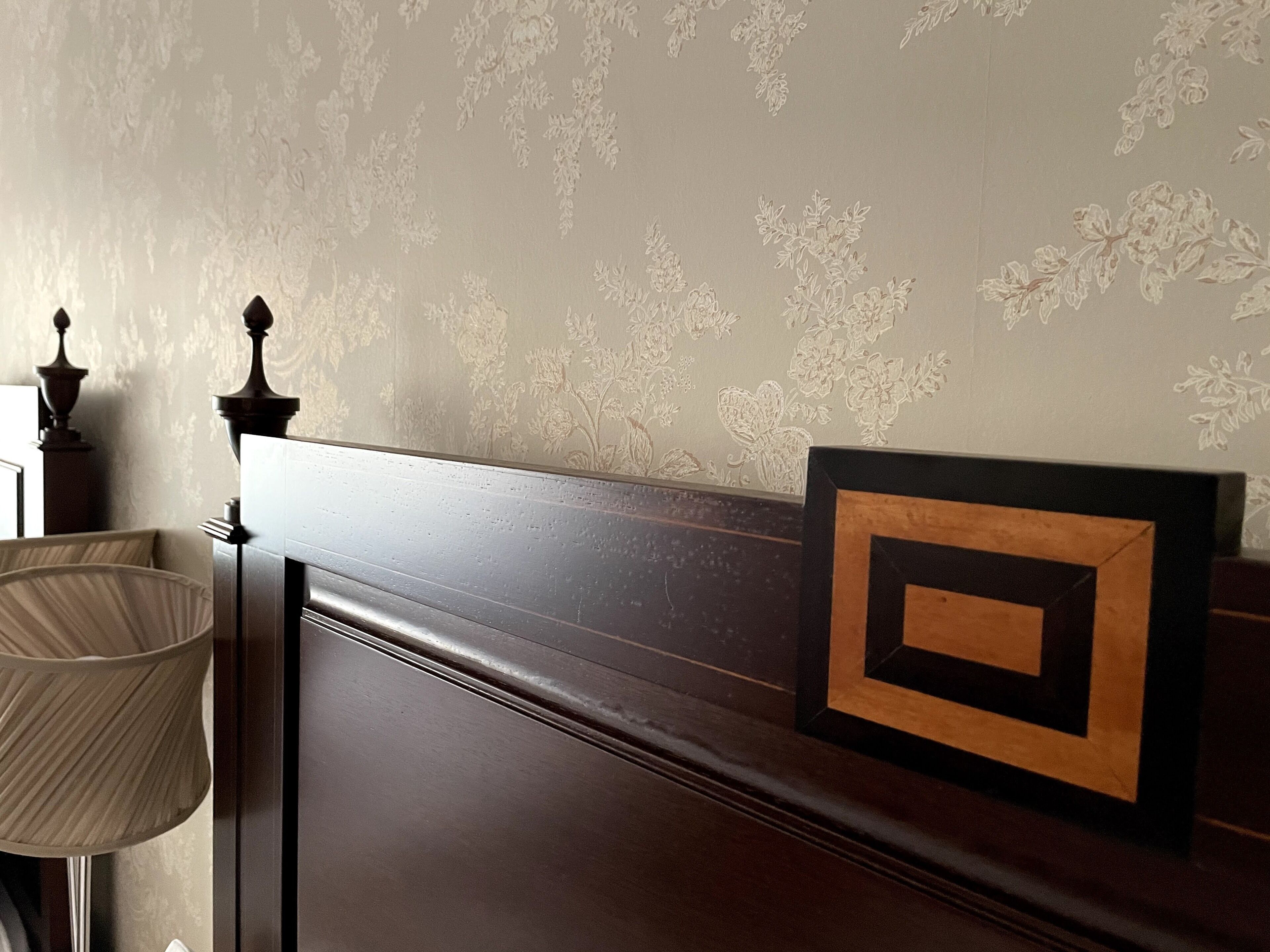 Een slaapkamer met een donker houten hoofdeinde, een bedlampje en behang met een bloemendesign.