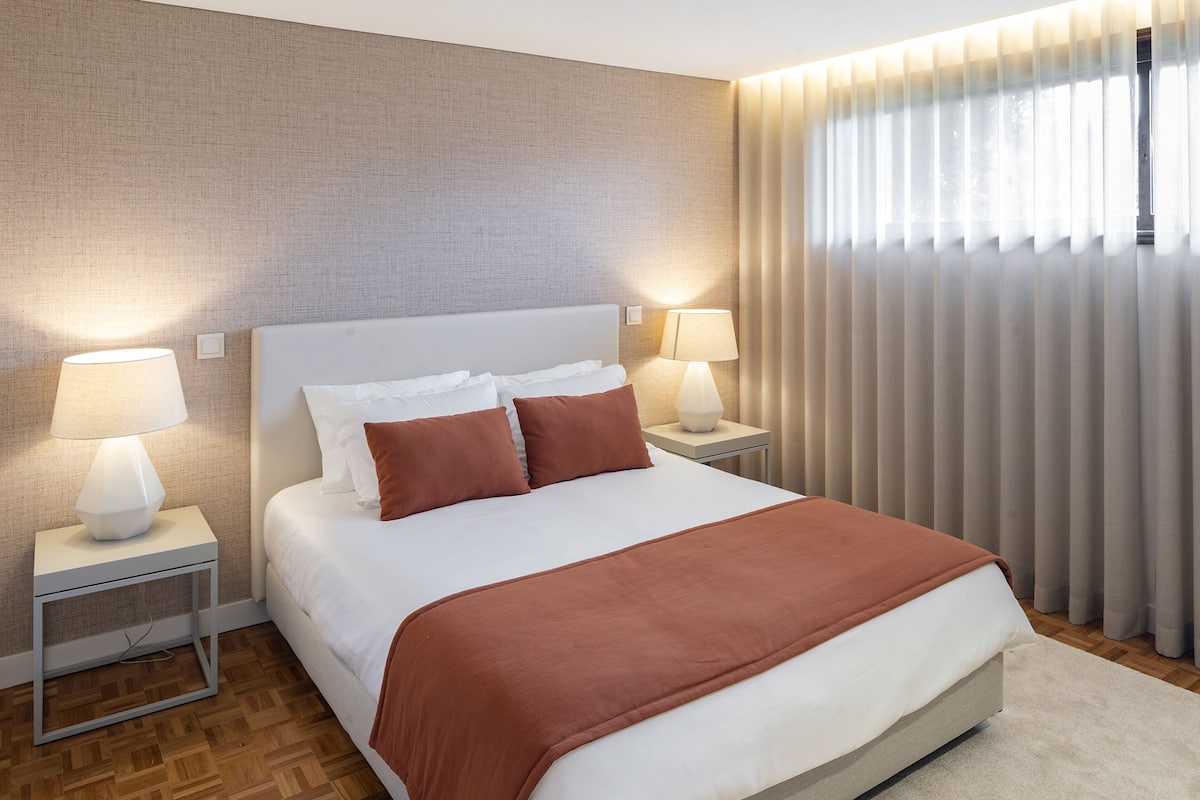 Een moderne hotelkamer met een groot bed, twee nachtkastjes met lampen, een hoofdbord, en uitzicht op het landschap door dunne gordijnen.