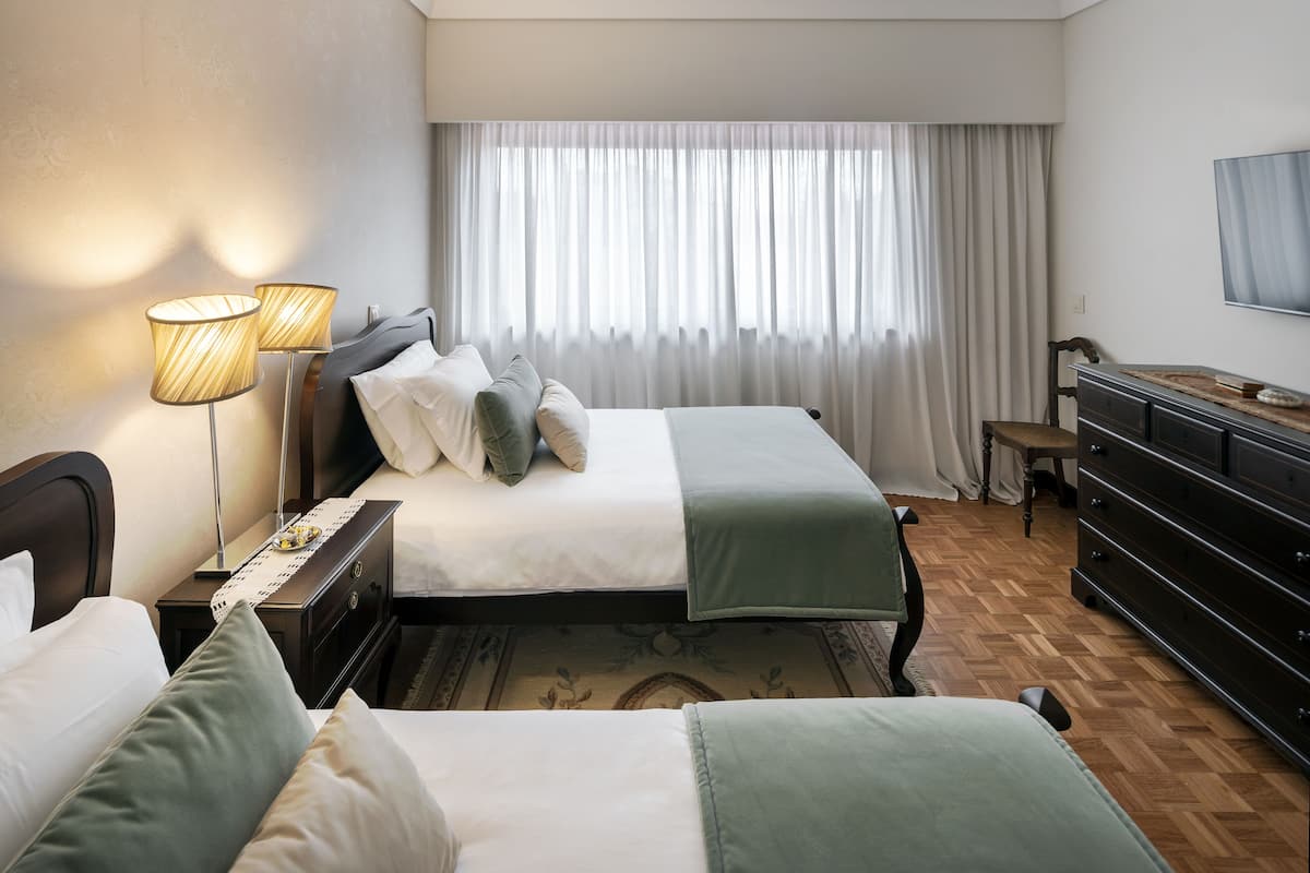 Een hotelkamer met twee bedden, een dressoir, een bureau met lamp en een groot raam met gordijnen.