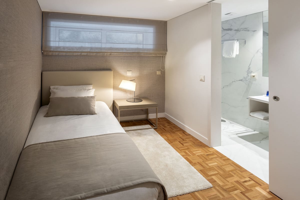 Een moderne hotelkamer met een bed, nachtkastje met lamp, klein nachtkastje, houten vloer en een douchecabine met glazen wanden.