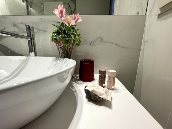 Un baño con un lavamanos grande de color blanco, una vela roja y dos pequeños frascos de productos Calimero sobre el mostrador.