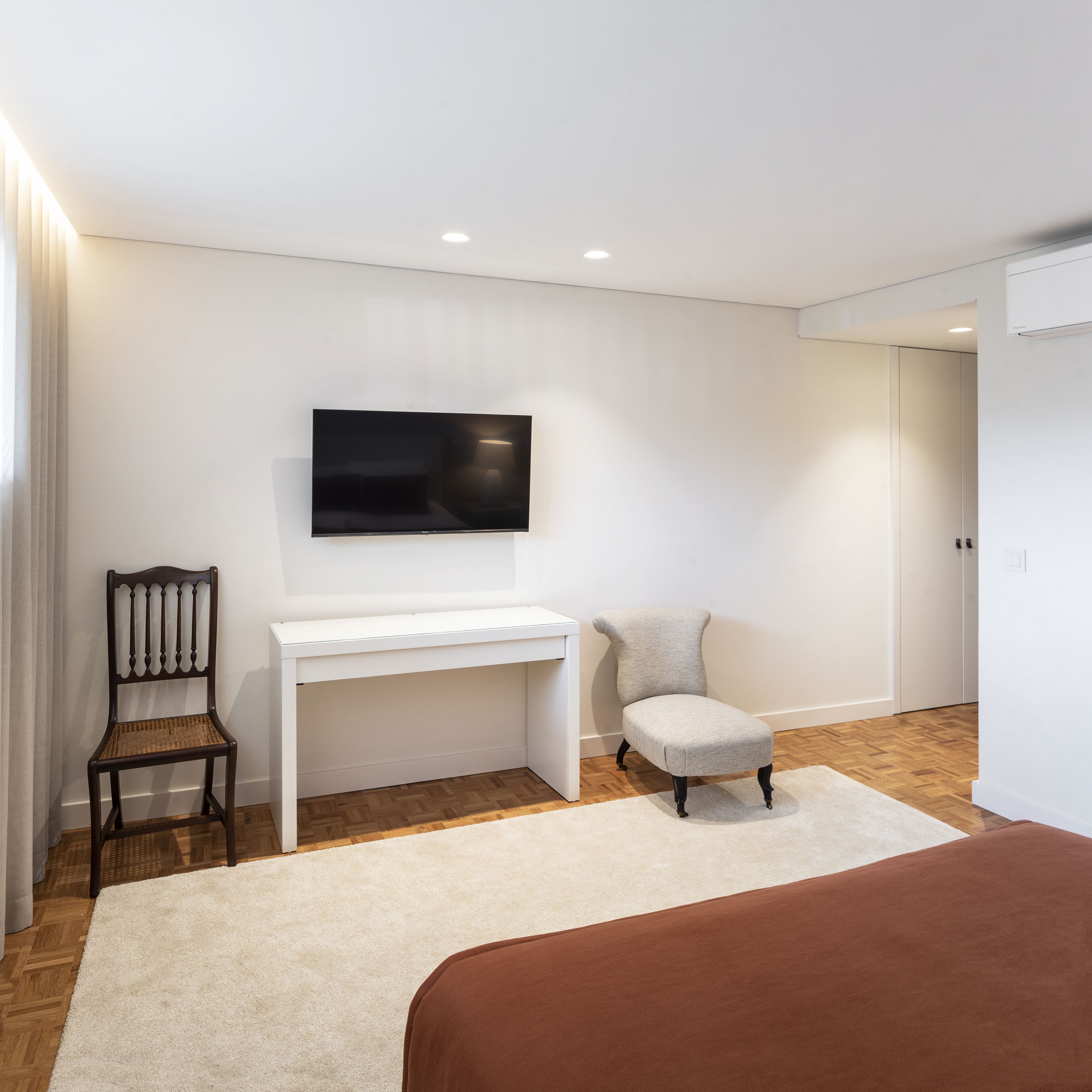 A modern bedroom with a white desk, a television mounted on the wall, a wooden chair, a brown ottoman, and a large bed with a brown bedspread.